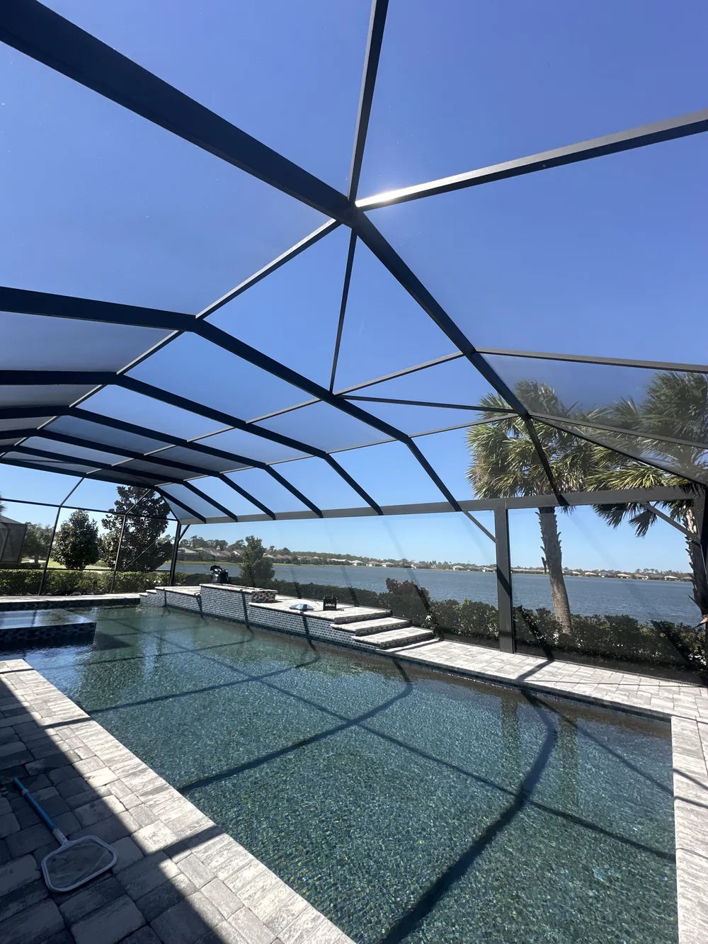 Inside a bronze aluminum pool cage with a panoramic screen section, looking across the pool to a lake and palm trees